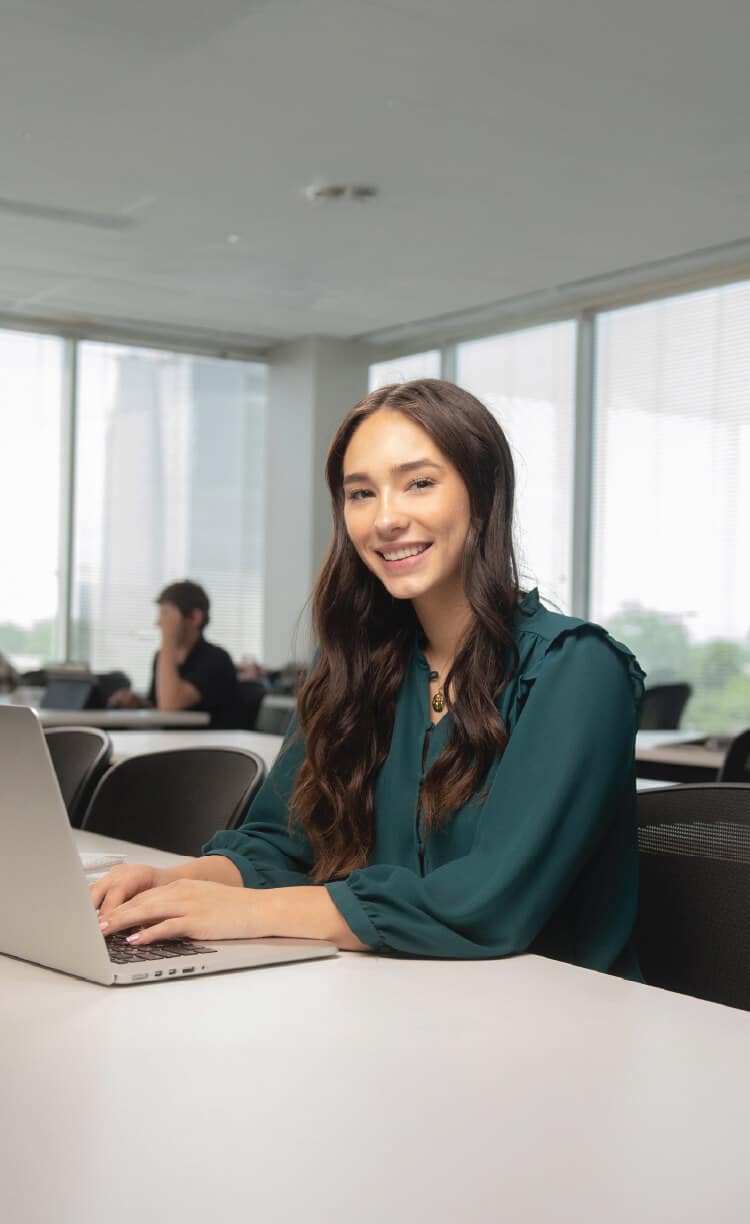 A young woman with long brown hair, wearing a teal blouse, sits at a white table using a laptop and smiling in a bright, modern classroom with large windows.