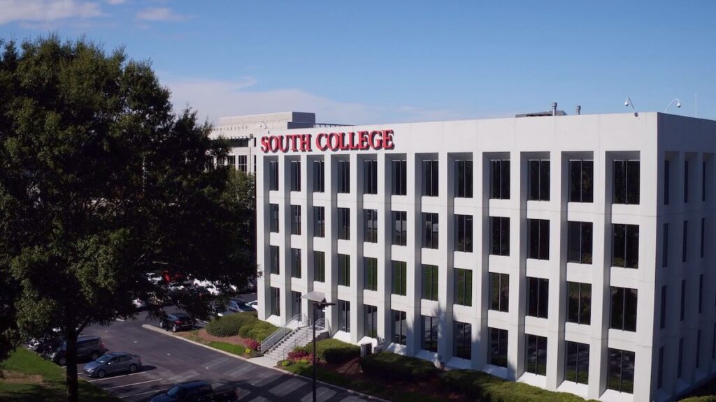 A four-story, white modern building with large, rectangular windows. "South College" is displayed in red letters near the top. The sky is clear and blue, and green foliage and parked cars can be seen around the building.
