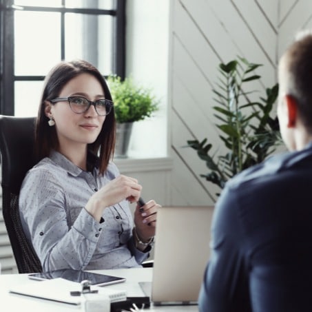 A woman wearing glasses sits at a desk with a laptop, facing a man. She is smiling and appears engaged in conversation. A large window and green plants are in the background, creating a bright and pleasant office environment.