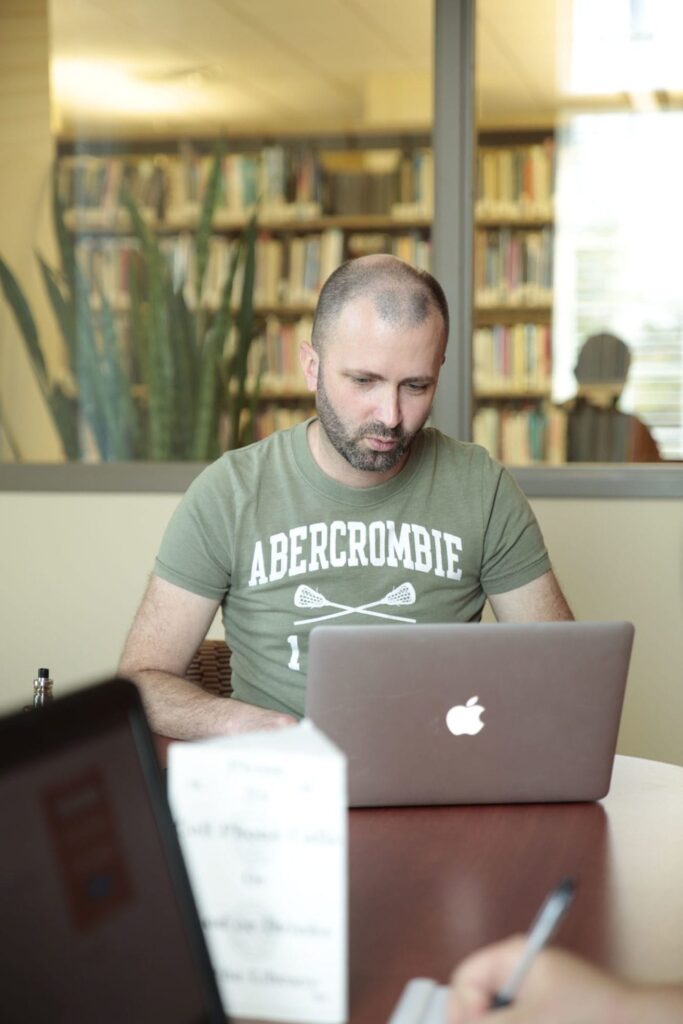 A man in a green "Abercrombie" shirt focuses intently on his laptop in a library setting.
