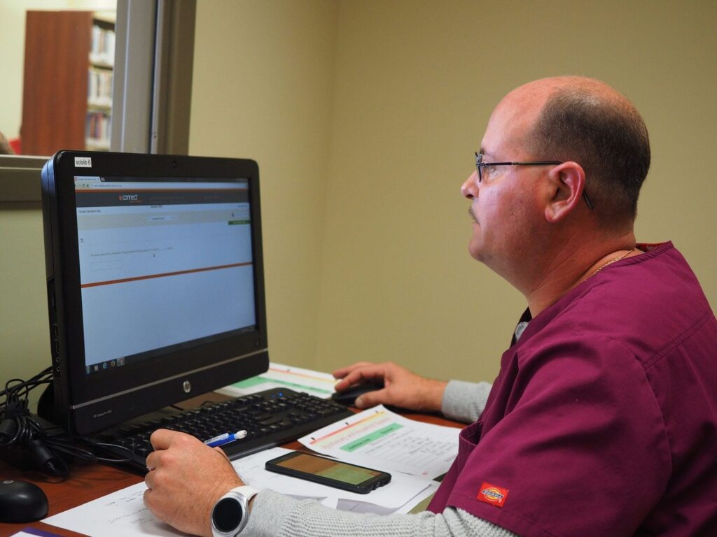 A nursing student in a maroon scrub top is sitting at a desk, focused on the computer screen.