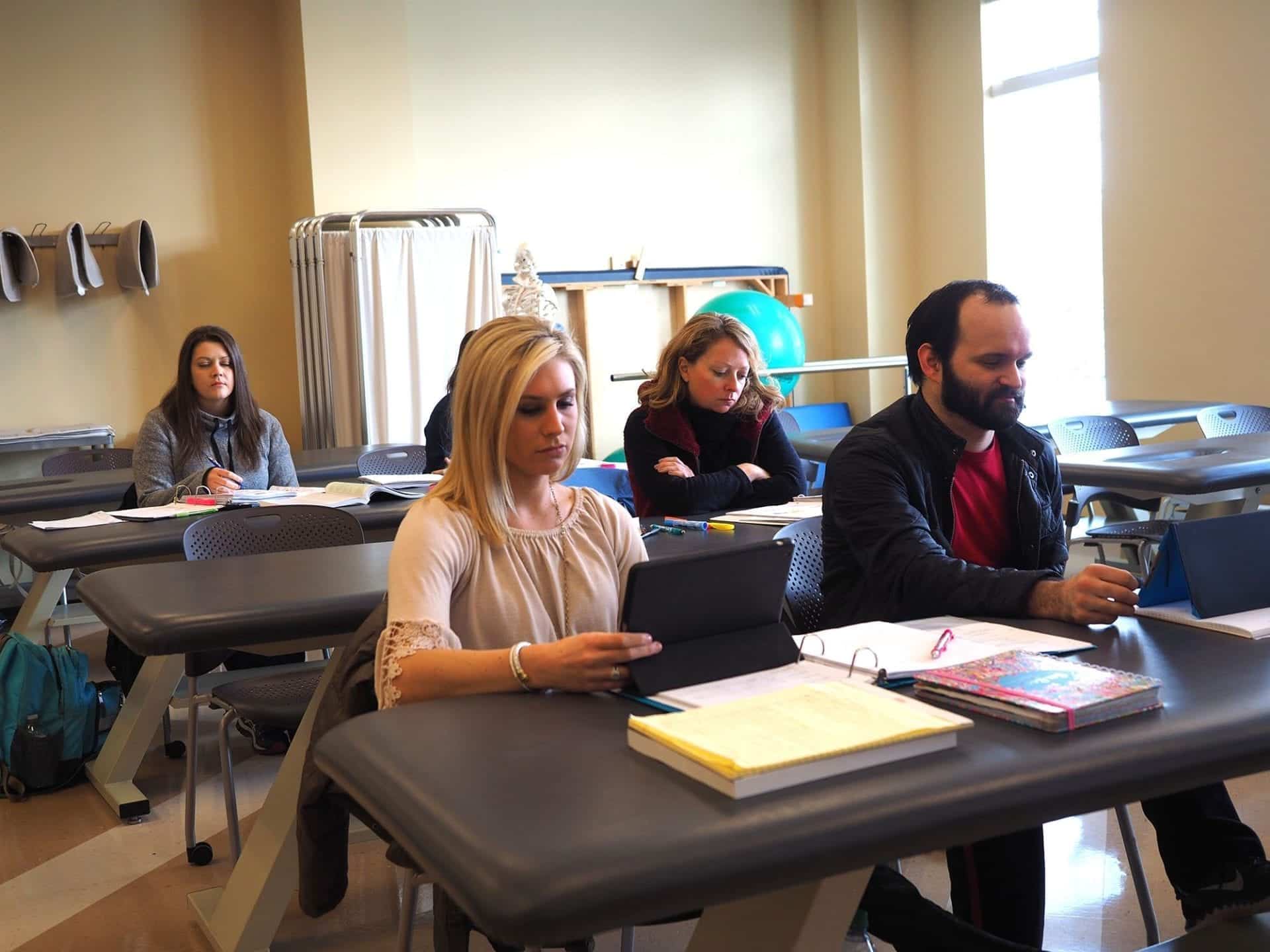 In a classroom, five students are seated at tables, attentively engaging in tasks.