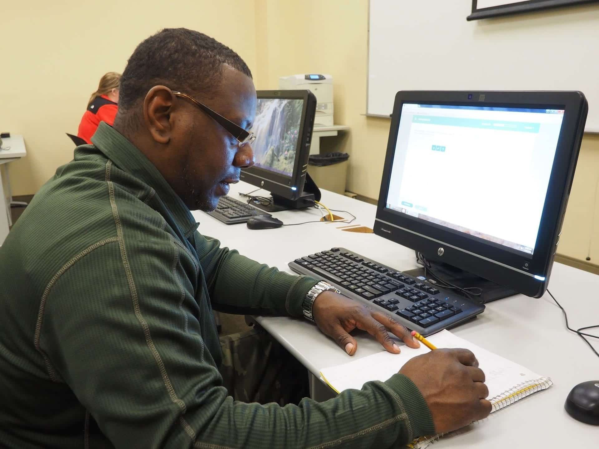 A student in a green jacket sits at a desk in a computer lab, jotting down notes in a notebook while glancing at a computer screen.