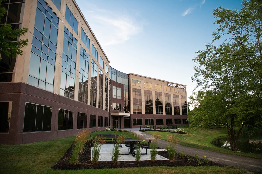 A modern, multi-story office building with large reflective windows and "South College" signage. The building is surrounded by well-maintained greenery, including trees and small plants. In the foreground, there is an outdoor sitting area with tables and chairs.