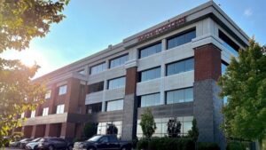 A four-story brick and glass building with trees in the foreground and cars parked in front. The building has the sign "DOHERTY COLLEGE" on the top. Sunlight is shining through the leaves of the trees.