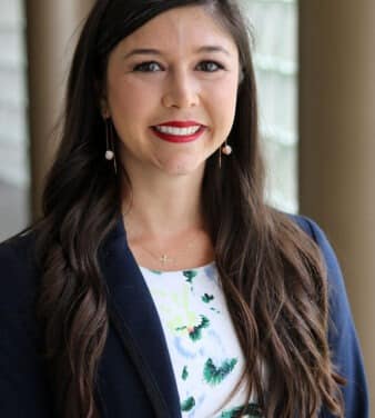A woman with long brown hair, wearing a white floral dress and a dark blazer, is smiling in front of a blurred background with large windows and columns. She is also wearing drop earrings and a delicate necklace.