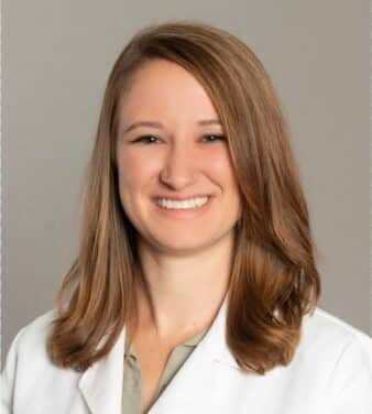 A smiling woman with shoulder-length light brown hair is wearing a white medical coat. The background is plain and neutral. She appears to be a healthcare professional.