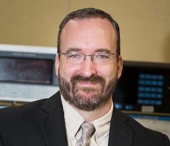 A man with short dark hair, glasses, and a beard smiles at the camera. He is wearing a dark suit with a light colored shirt and tie. The background shows electronic equipment with screens and buttons.