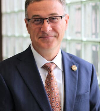 A man wearing glasses, a dark pinstripe suit jacket, a white dress shirt, and an ornate patterned tie stands in front of a frosted glass window. He is smiling slightly and has short hair. A pin with a South College emblem is attached to his jacket lapel.