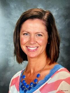 A woman with shoulder-length brown hair smiles at the camera. She is wearing a striped top in shades of pink, white, and blue, and a blue necklace with pendant details. The background is a soft, blended gray.