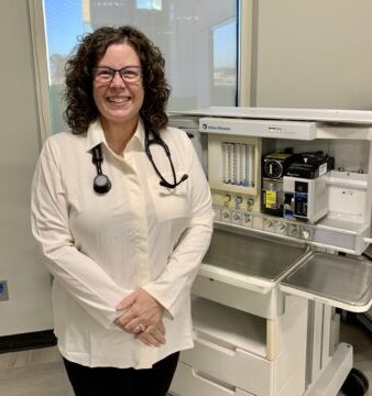 A smiling woman with curly hair and glasses, wearing a white blouse and a stethoscope around her neck, stands next to a medical machine in a clinical setting.