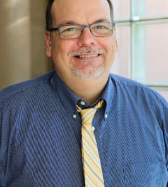 A man wearing glasses, a blue striped shirt, and a yellow tie is smiling at the camera. He has short hair, a goatee, and is standing in front of a window with sunlight streaming in, creating a bright and warm background.