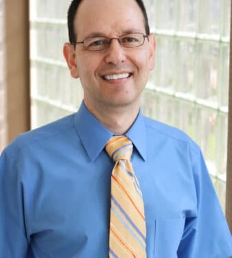 A man with short dark hair and glasses smiles while wearing a blue dress shirt and a yellow striped tie. He stands in front of a wall with glass blocks.