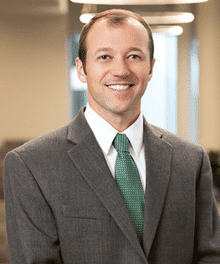 A man wearing a gray suit, white dress shirt, and green tie is smiling. He has short brown hair and stands in an office setting with blurred background elements including windows and lights.