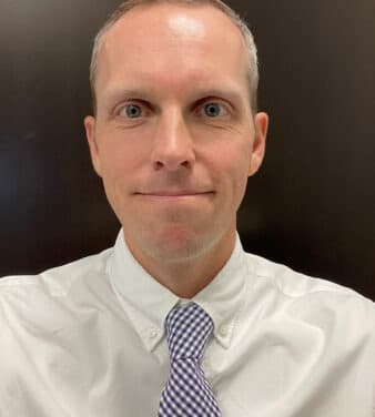 A man with short blonde hair and blue eyes is wearing a white collared shirt and a blue and white checkered tie. He is smiling slightly and standing in front of a dark background.