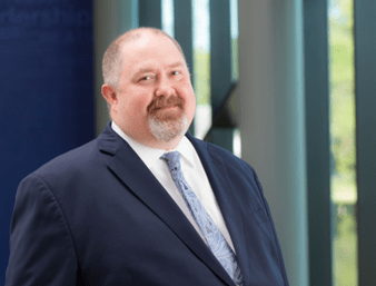 A man with a beard and mustache, wearing a navy suit, white dress shirt, and patterned tie, stands in front of a blurred background with vertical windows. He has a slight smile and a professional appearance.