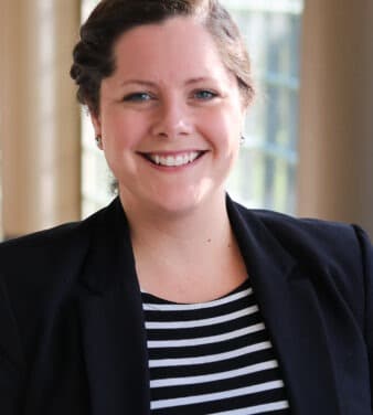 A woman with brown hair pulled back, wearing a black blazer and a black and white striped top, is smiling at the camera. She is standing in front of a light background with large windows.