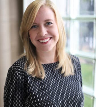 A woman with shoulder-length blonde hair is smiling and looking at the camera. She is wearing a black top with white patterns and is standing indoors next to a large, light-filled window with glass blocks.