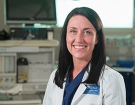 A smiling woman with long dark hair, dressed in a white doctor's coat over navy scrubs, stands in a medical setting featuring various equipment and monitors in the background.