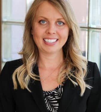 A woman with long, wavy blonde hair and blue eyes is smiling at the camera. She is wearing a black blazer over a black and white patterned top. The background features a window with panes.