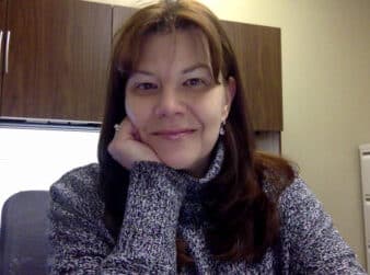 A woman with long brown hair and wearing a gray sweater is sitting at a desk with her head resting on her hand. She is smiling and looking directly at the camera. The background shows a wooden cabinet and part of a filing cabinet.