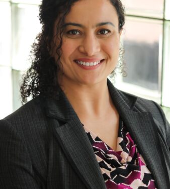 A person with curly dark hair smiles at the camera. They are wearing a black blazer over a patterned blouse, standing in front of a window with natural light filtering in.