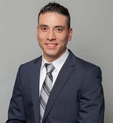A man with short, dark hair is smiling while wearing a dark blue suit, a white shirt, and a striped tie. He is posed against a plain, light grey background.