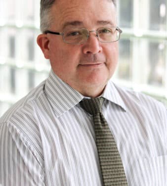 A man with short hair, glasses, and a light striped shirt with a dark striped tie stands indoors, posing in front of a backdrop of glass blocks. He has a neutral expression on his face.