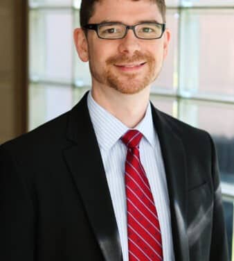 A man wearing glasses, a dark suit jacket, a light blue dress shirt, and a red striped tie stands smiling in front of a sunlit window.