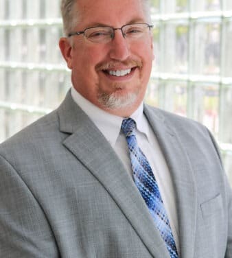 A smiling man with short hair and glasses is wearing a gray suit, white shirt, and blue patterned tie. He is standing in front of a backdrop made of glass bricks.
