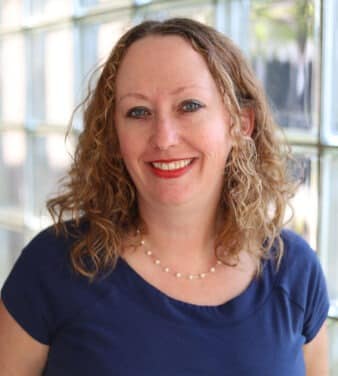 A woman with curly, shoulder-length hair wearing a navy blue shirt and a pearl necklace is smiling at the camera. She is standing in front of a wall made of glass blocks, with natural light illuminating the scene.