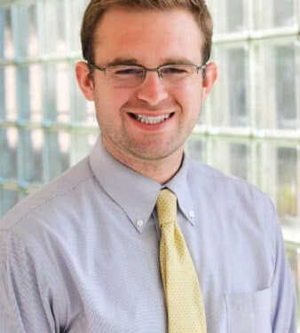 A man with short brown hair, glasses, and a trimmed beard is smiling. He is wearing a light blue dress shirt and a yellow tie. The background features a wall of glass blocks, softly lit from behind.