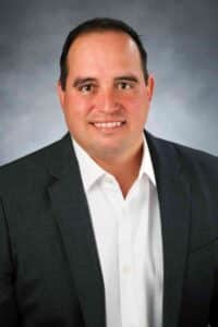 A man with short dark hair is smiling and wearing a dark grey blazer over a white dress shirt. The background is a gradient of grey tones.