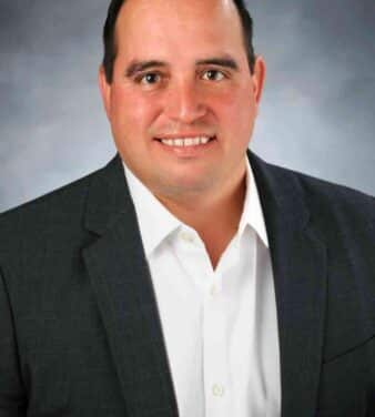 A man with short dark hair is smiling and wearing a dark grey blazer over a white dress shirt. The background is a gradient of grey tones.