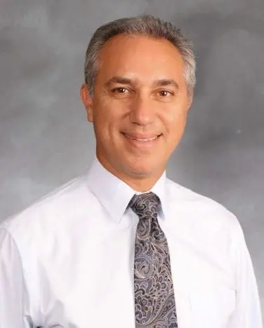 A man with short hair wearing a white dress shirt and a patterned tie smiles in front of a grey background.