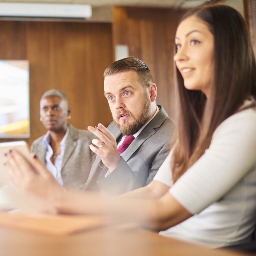 Three individuals are sitting at a conference table. A man in a suit and tie appears to be speaking and gesturing with his hand. A woman in a white top is sitting beside him, attentively listening. Another man in a casual blazer sits in the background.