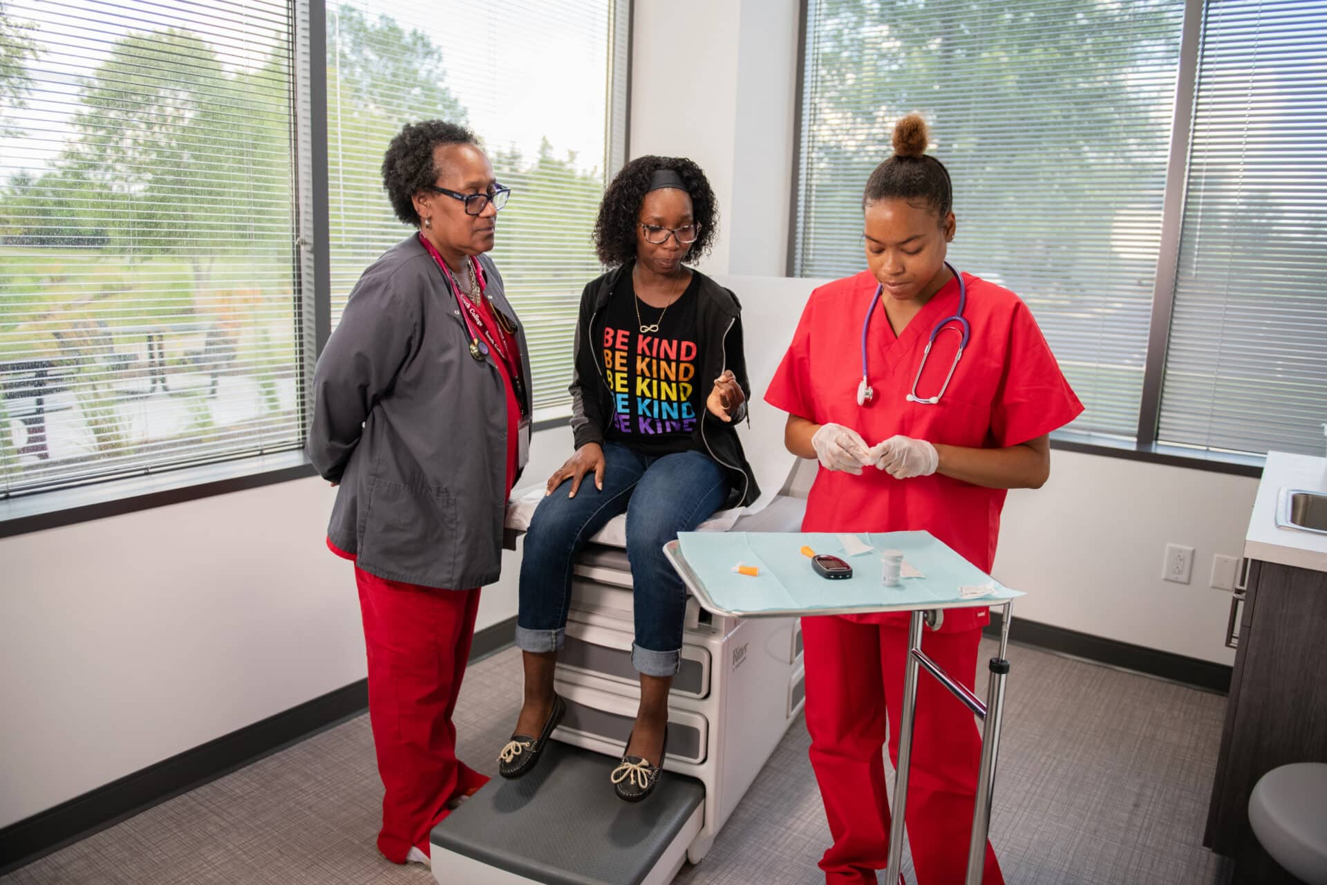 Two medical assisting students in red scrubs assist a seated patient in a medical exam room. The patient wears casual clothing and a colorful shirt with positive messages. One professional prepares medical supplies on a tray, while the other looks on.