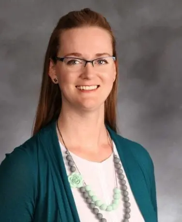 A woman with long brown hair and glasses is smiling at the camera. She is wearing a teal cardigan over a white top accessorized with a necklace of grey and mint green beads. The background is a grey, marbled texture.