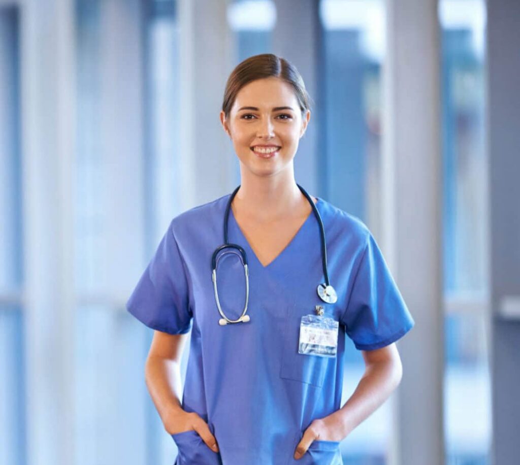 A medical assisting student wearing blue scrubs with a stethoscope around their neck stands in a well-lit medical facility. They have their hands in their pockets and are smiling at the camera. Their ID badge is visible, and large windows are in the background.