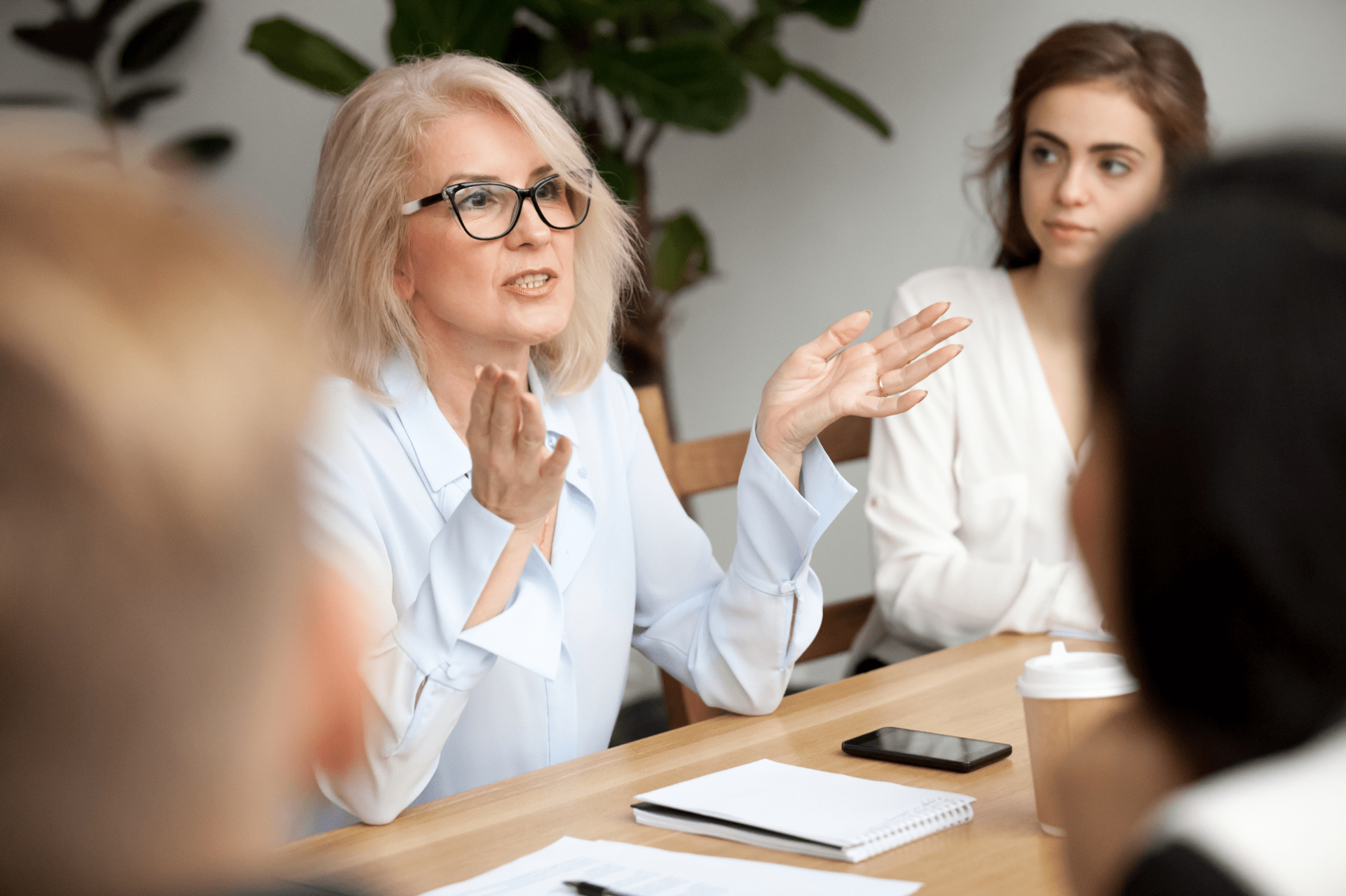 A woman with glasses and short blonde hair is speaking in a meeting. She is seated at a wooden table with a notebook, a smartphone, and a coffee cup in front of her. Other attendees, including a woman with long brown hair, are listening attentively.