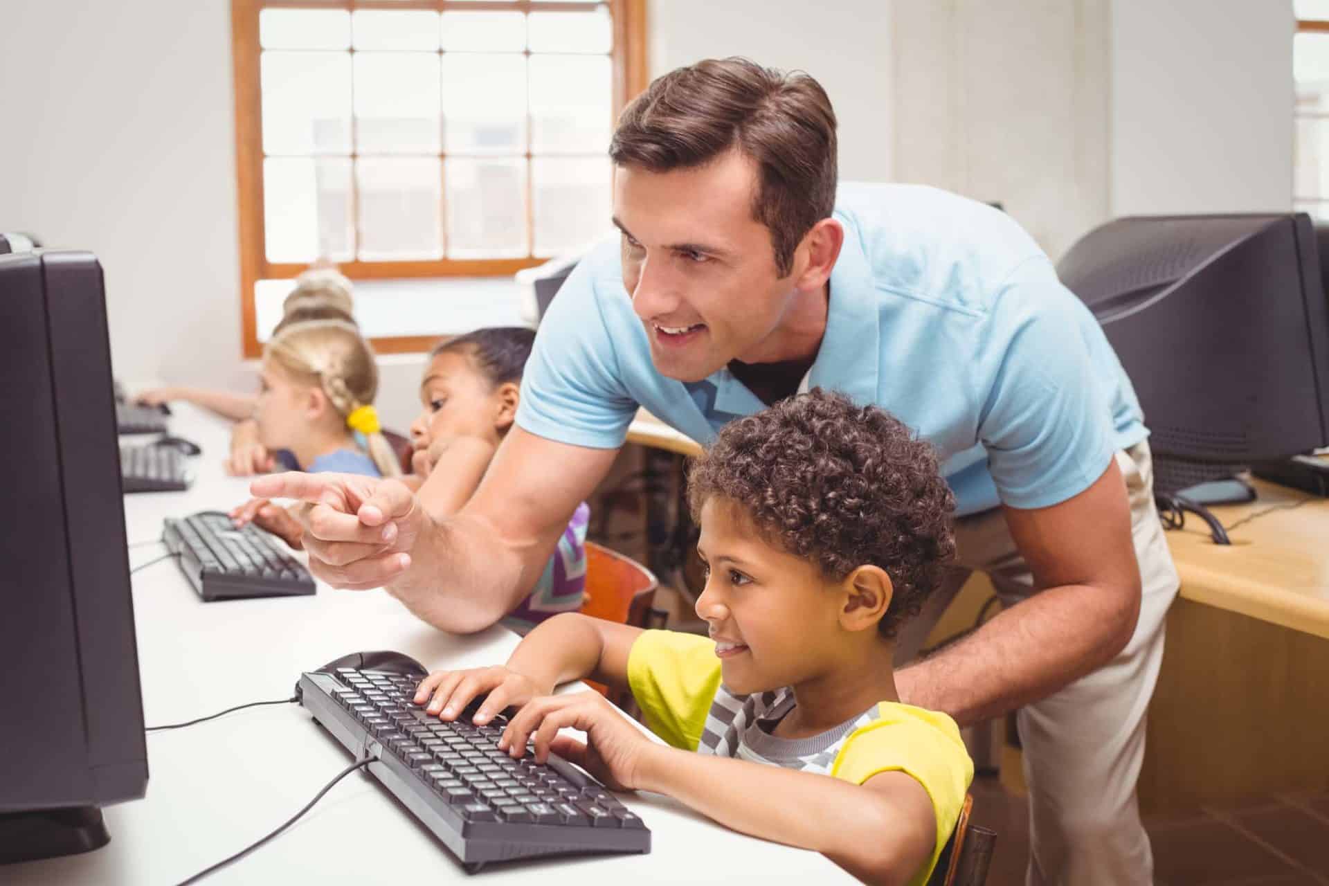 A teacher is helping a smiling student who is using a desktop computer in a classroom. Other children are also working on computers in the background. The classroom is well-lit with natural light coming through the windows.