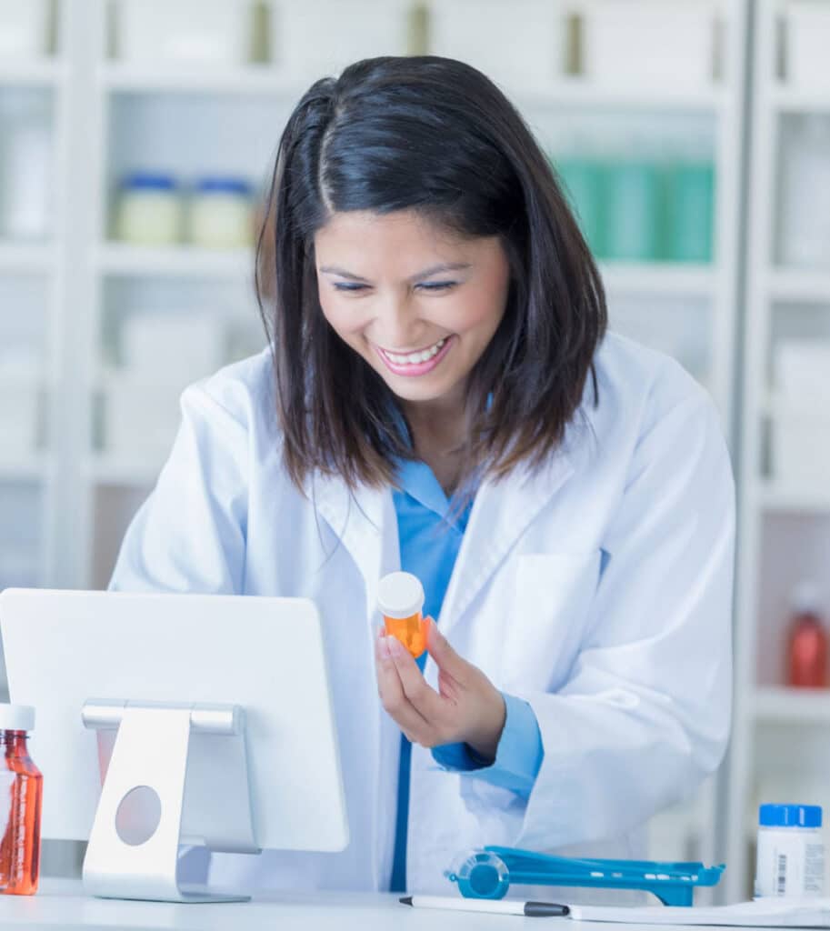 A female pharmacist in a white lab coat is holding a prescription bottle and smiling while looking at a computer screen in a pharmacy. Shelves with various bottles and containers are visible in the background.