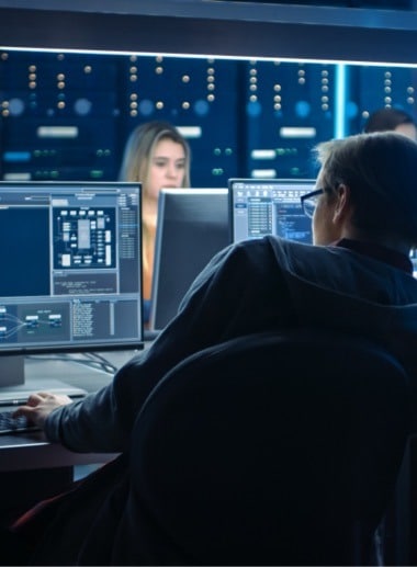 A person in glasses sits at a desk with multiple computer monitors, focusing intently on the screens. The background shows another individual working at a similar setup, with servers or mainframes visible behind them. The room is dimly lit with blue lighting.