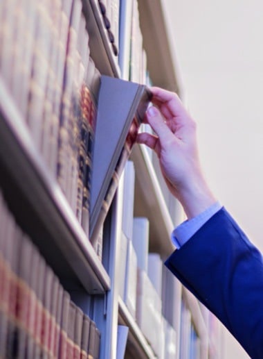 A hand in a blue suit jacket reaches out to pull a book from a tall bookshelf filled with various books in an organized library setting.