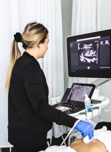 A student with long blonde hair in a ponytail is wearing a black jacket and blue gloves while using an ultrasound probe on a patient's abdomen. They are looking at a monitor displaying ultrasound images. The room has white curtains in the background.