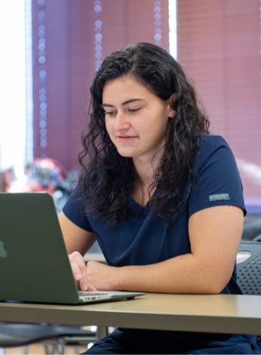 A person with long, dark curly hair wearing a navy blue top is sitting at a desk using a laptop. They are indoors, and light is coming through a window with closed blinds in the background.