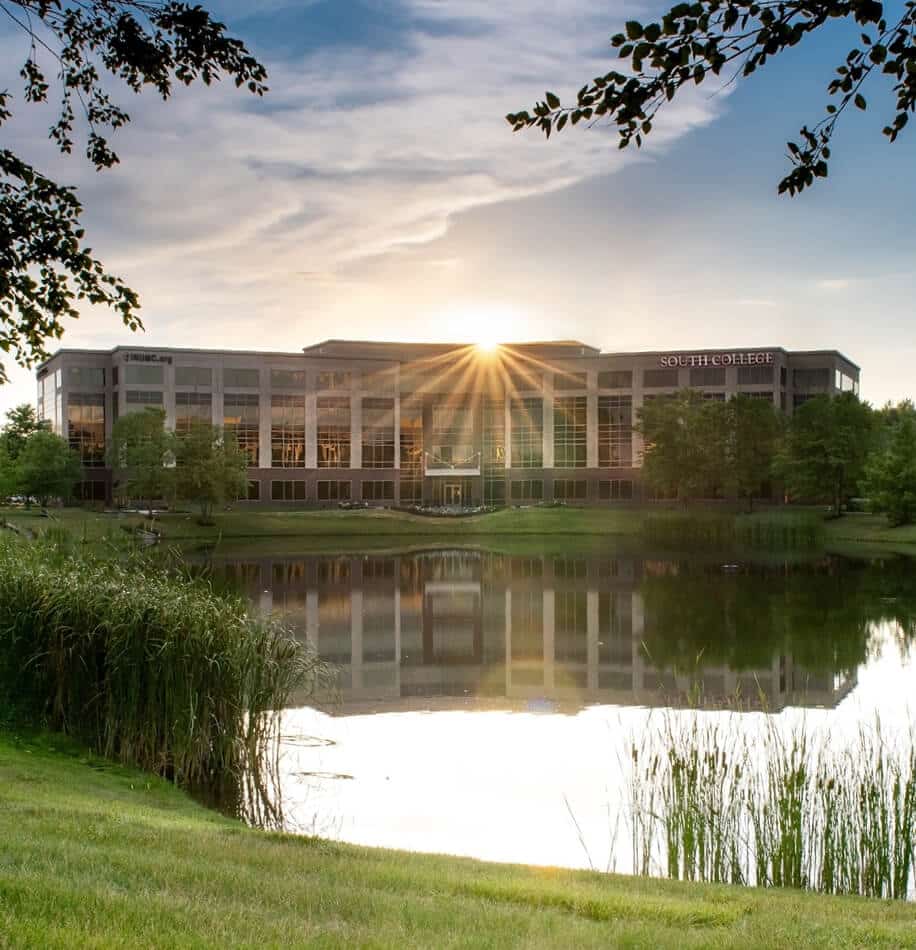 A large modern building with the text "South College" on it sits by a tranquil pond surrounded by green grass and trees.