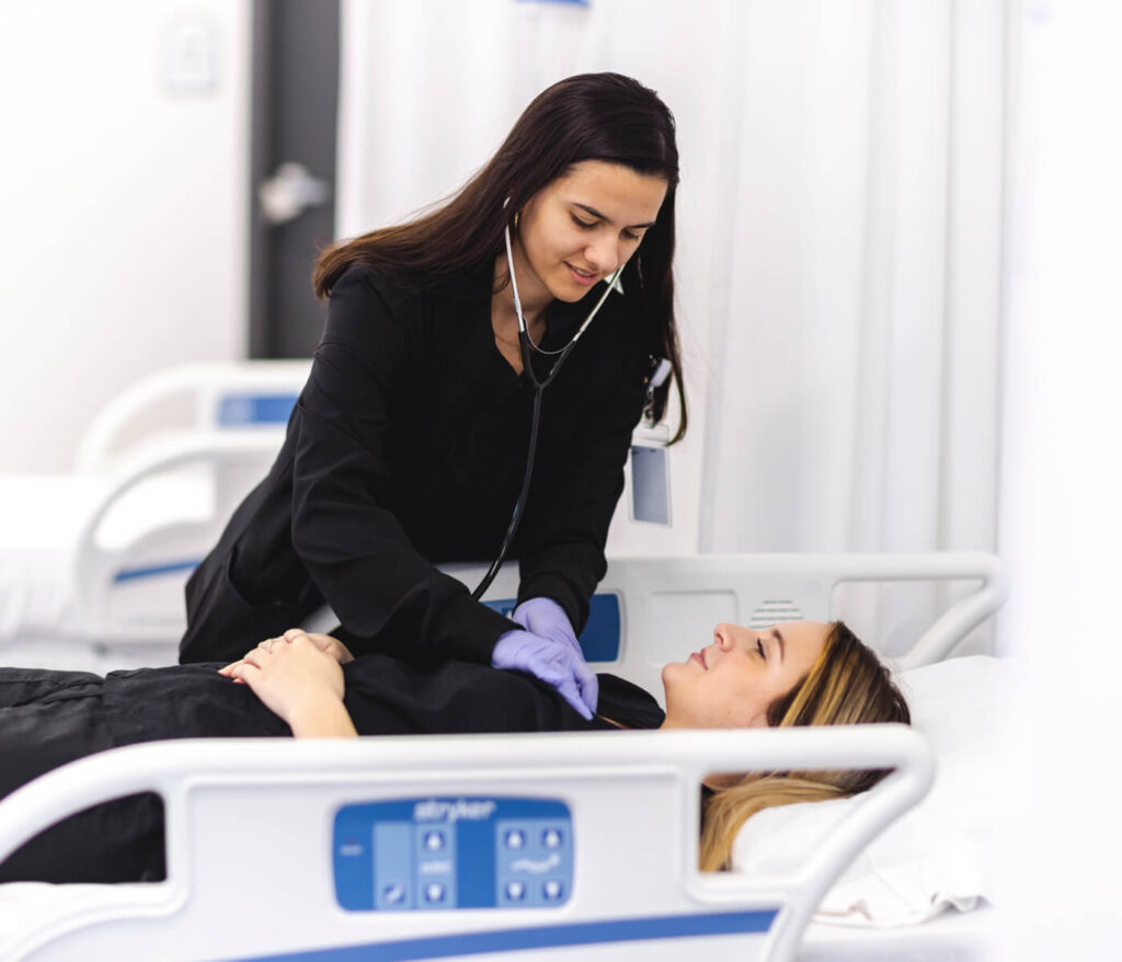 A nursing student in a black coat and purple gloves uses a stethoscope to listen to the heart of a patient lying in a hospital bed.
