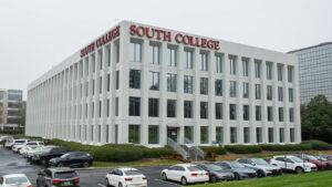 A modern white building with large windows and red "South College" signs is surrounded by a parking lot filled with cars.