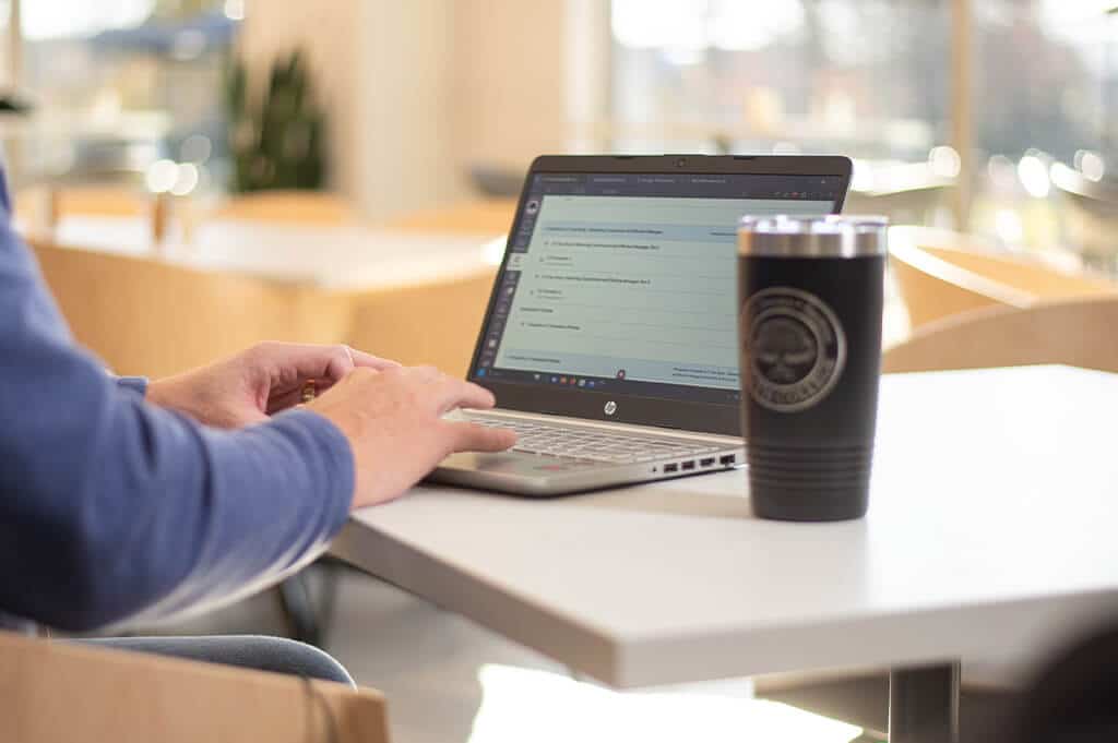 A person in a blue sweater is typing on a laptop at a white table in a brightly lit cafe.